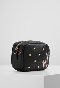 Musta tikattu meikkilaukku kultaisilla sydänkoristeilla ja huulipainatuksella. Siinä on vetoketjullinen suljin ja suorakulmainen muoto. Sileä tekonahkapinta.