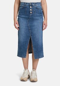 Jupe midi en jean de couleur bleu clair avec une fente à l'avant et une fermeture à cinq boutons. Dotée de passants de ceinture et d'un design légèrement décoloré. Portée avec des baskets beiges.