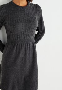 Femme portant une robe à manches longues en maille gris foncé avec un motif côtelé et torsadé sur la moitié supérieure, sur un fond clair uni.