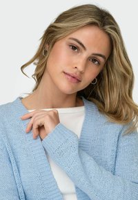 Young woman with wavy blonde hair wearing silver hoop earrings, a white top, and a light blue knitted cardigan against a plain background.