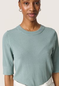 Ljus teal-färgad stickad T-shirt med ribbad textur, rund halsringning och halvlånga ärmar. Tyget har små vertikala öppningar för ventilation.