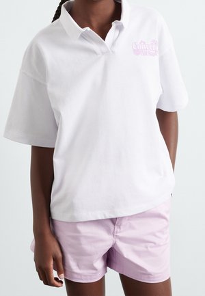 Personne portant un polo blanc à manches courtes avec le logo « Converse All Star » et un short violet clair, debout devant un fond uni.