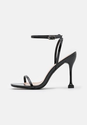 Blank sort sandal med høj hæl med tynde stropper over tæerne og anklen, med en lille spænde og en slank, buet hæl.