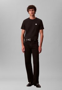Mustast puuvillast T-särk väikese logoga, millele on sobitatud mustad sirge lõikega teksapüksid ja must vöö. Modell seisab neutraalse tausta ees.