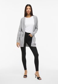 Grauer langer Cardigan mit Taschen, getragen über einem weißen Hemd, kombiniert mit schwarzen Leggings und offenen schwarzen High Heels. Einfache, leichte Strickstruktur.