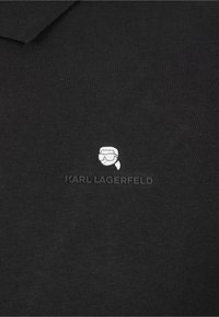 Svart bomullspolo med ribbad krage, försedd med en vit tryckt logotyp av en karaktär och texten "KARL LAGERFELD" under.