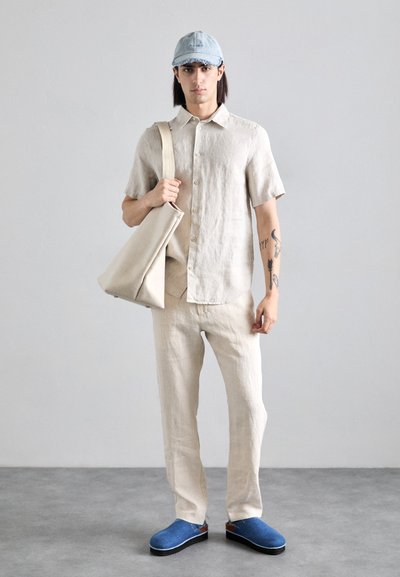 Lichtbeige linnen shirt, bijpassende beige broek, blauwe klompen en een denim pet. Model houdt een beige tote bag vast met een minimalistisch ontwerp.