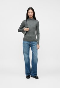 Kvinde iført en grå ribbet langærmet top, brede blå jeans, sorte sko og holder en lille mønstret skuldertaske på hvid baggrund.