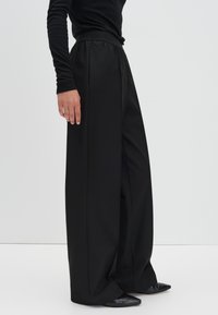 Pantalon large noir avec une taille élastique haute, présentant des plis subtils, un tissu lisse et un design épuré et minimaliste.