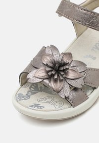 Metallisk grå sandal med blomstermønster på remmen, med teksturerte kronblader og et mønstret såle med sommerfugler.