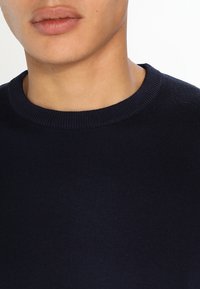 Mörkblå stickad tröja med ribbat rundhals. Mjuk textur och figurnära design. Synlig hud på halsen och l äpparna i förgrunden.