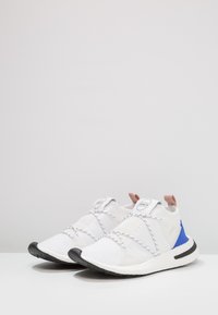 Valkoiset urheilukengät, joissa on verkkopäällinen, musta kumipohja ja siniset korostukset. Varustettu joustavilla nauhoilla ja pehmustetulla kantapääsysteemillä.