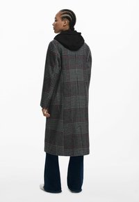 Abrigo largo gris con un patrón de cuadros, con una capucha negra, corte recto y bolsillos. Combinado con jeans acampanados azules.