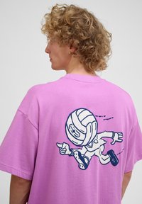 Lilla t-shirt lavet af bomuld, med en tegneseriefigur trykt i marineblå på ryggen, der skildrer en figur, der løber med en bold.
