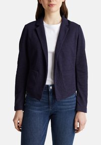 Blazer azul marino con solapas de muesca, dos bolsillos delanteros y tejido texturizado. Lleva una camiseta blanca debajo, combinado con jeans azules.