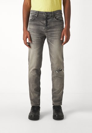 Mann trägt ausgewaschene graue Jeans mit Riss am rechten Knie, ein gelbes Shirt und schwarze Sneakers, steht vor weißem Hintergrund.
