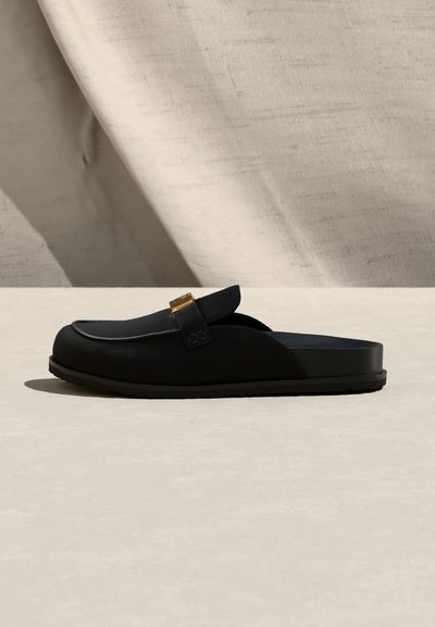Μαύρο μοκασίνι slip-on με χοντρή σόλα και λεπτομέρεια με χρυσή αγκράφα, τοποθετημένο σε μπεζ υφασμάτινο φόντο με απαλό φωτισμό.