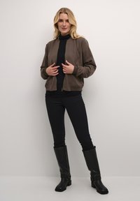 Veste en cuir marron avec des poignets et une taille côtelés, assortie à un col roulé noir, un jean skinny noir et des bottes noires jusqu'au genou.