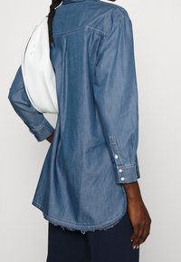 Spijkerblouse in middenblauw met lange mouwen, knoopmanchetten en een gebogen zoom. Een wit crossbodytasje hangt over de schouder.