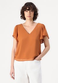 Top con escote en V en tejido ligero de color óxido. Presenta mangas cortas con volantes y un corte holgado. Combinado con pantalones blancos.