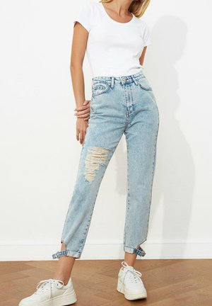 Frau trägt hellblaue, hoch taillierte, zerrissene Jeans mit Knöchelriemen, weißes, kurzärmeliges Crop-Top und weiße Plateau-Sneaker, steht drinnen.