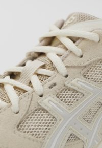 Beige sportskor med nät- och mockamaterial, platta snören och en framträdande vit logotyp på sidan. Texturerad yta synlig.