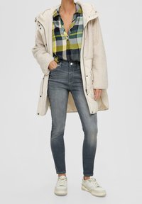 Beige, fluffy lang frakke over en grøn og blå rutet knap-op skjorte. Fadede grå skinny jeans og hvide sneakers med beige accenter.