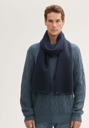 Una bufanda de lana navy se desliza alrededor del cuello, presentando un diseño limpio. El suéter de punto tiene una textura que resalta un patrón de hojas en tonos azules.