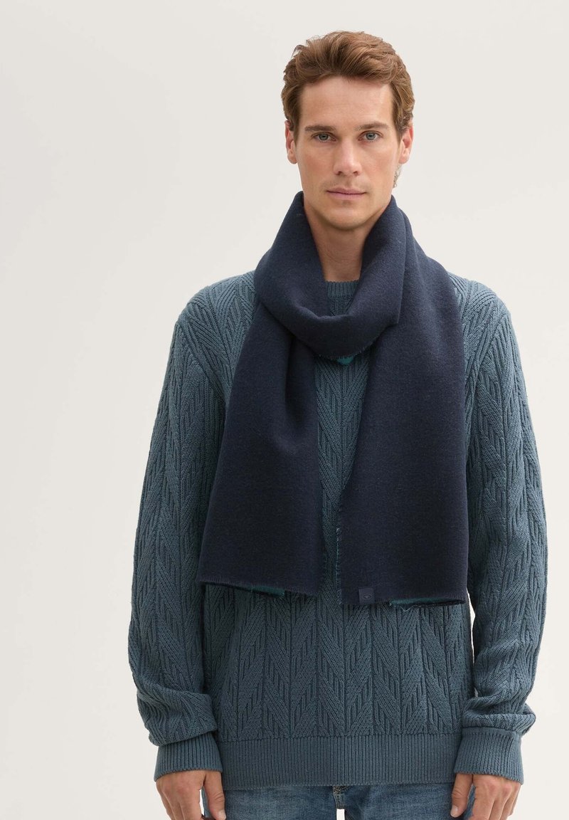 Una bufanda de lana navy se desliza alrededor del cuello, presentando un diseño limpio. El suéter de punto tiene una textura que resalta un patrón de hojas en tonos azules.