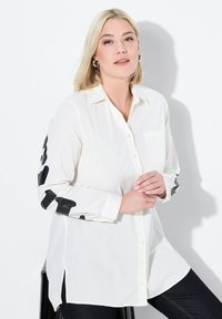 Camicia bianca a maniche lunghe con macchie astratte nere sulle maniche, bottoni frontali e taschino sul petto. Tessuto morbido con una vestibilità comoda.