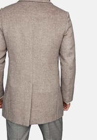 Cappotto a spina di pesce marrone con una vestibilità sagomata, caratterizzato da un colletto a rever e una spacco singolo sul retro. Abbinato a pantaloni a quadri.