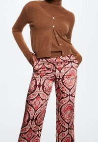 Cardigan marron à boutons en maille douce, porté avec un pantalon large présentant un motif paisley rouge et crème sur fond sombre.