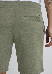 Shorts en coton vert olive à texture douce, dotés d'une poche arrière et d'une coupe décontractée, montrant des détails de coutures et une finition à l'ourlet.