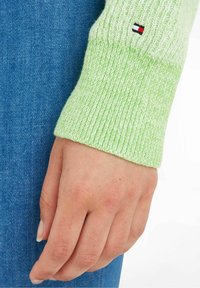 Hand liegt seitlich und trägt einen hellgrünen Strickpullover mit einem kleinen rot-weißen und schwarzen Logo-Patch, vor einem blauen Denim-Hintergrund.