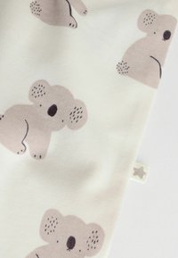 Bomuldsstof med en lys cremefarvet baggrund og gentagne beige koala mønstre. Blød tekstur med sorte detaljer på koalaerne.