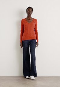 Orange knitted v-neck sweater with cable pattern, paired with dark blue flared jeans and white sneakers. Simple design, fitted silhouette.