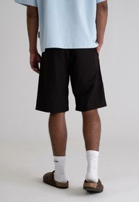 Black shorts made of soft fabric, featuring a relaxed fit and side pockets. Worn with brown sandals and white ribbed socks.