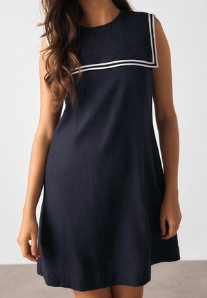 Femme portant une robe bleu marine sans manches avec des doubles rayures blanches sur le col carré, debout devant un fond clair uni.