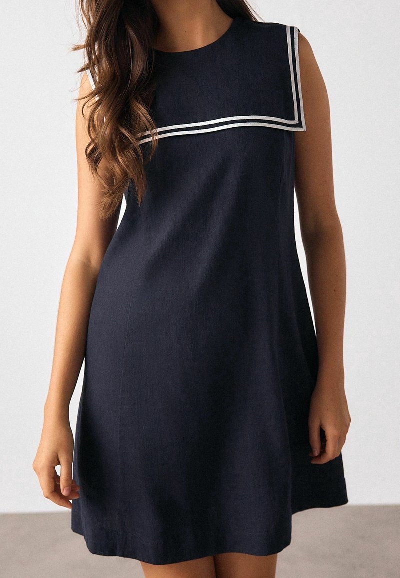 Femme portant une robe bleu marine sans manches avec des doubles rayures blanches sur le col carré, debout devant un fond clair uni.