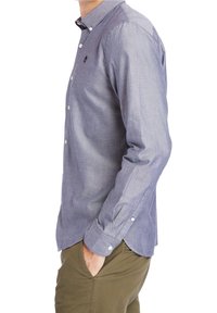 Uomo che indossa una camicia azzurro chiaro con bottoni e maniche lunghe, insieme a pantaloni verde oliva, in piedi con una mano in tasca, vista di lato.