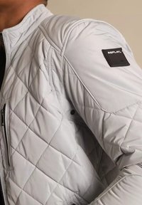 Giacca trapuntata grigia chiara con colletto rialzato, dotata di un patch con il logo REPLAY nero e una tasca con zip sul lato sinistro.