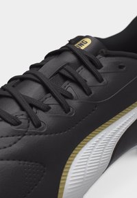 Chaussure de sport noire avec lacets noirs, accents dorés et semelle texturée blanche, vue en gros plan de côté et de dessus.