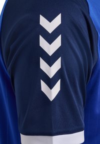 Blå sportstrøje lavet af syntetisk stof, med et hvidt chevronmønster på skulderen og en kontrastfuld hvid kant ved kanten.