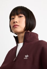 Sudadera burdeos con cremallera y cuello alto, que presenta un logo bordado de Adidas en blanco en el pecho y una tela suave y texturizada.