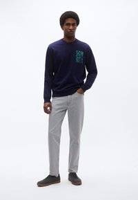 Tmavomodrá košeľa s dlhým rukávom s zeleným grafickým motívom na hrudi, v kombinácii so svetlosivými nohavicami a čiernymi teniskami, s voľným strihom.