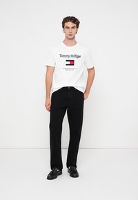 Ung mand iført en hvid Tommy Hilfiger t-shirt, sorte bukser og sorte sko, stående mod en hvid baggrund.