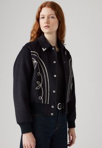 Melna vilnas bomber jaka ar baltām izšūtām ziedu dizainiem un sudraba pogām, ar ribotiem piedurknēm un apkakles izgriezumu.
