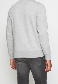 Person iført en lys grå langærmet sweatshirt og mørkegrå jeans set fra ryggen, stående mod en ensfarvet baggrund.