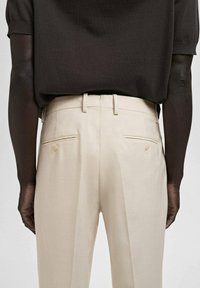 Beige maßgeschneiderte Hose mit glatter Textur, ausgestattet mit zwei Gesäßtaschen mit Knöpfen und einem Gürtelschlaufenbund, von hinten betrachtet.