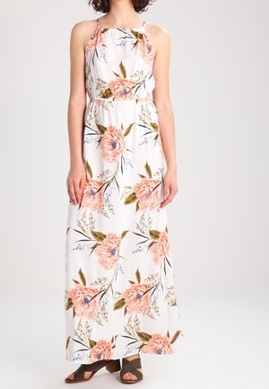 Woman wearing a sleeveless white maxi dress with pink floral patterns and olive green open-toe block heel sandals, standing against a plain background.